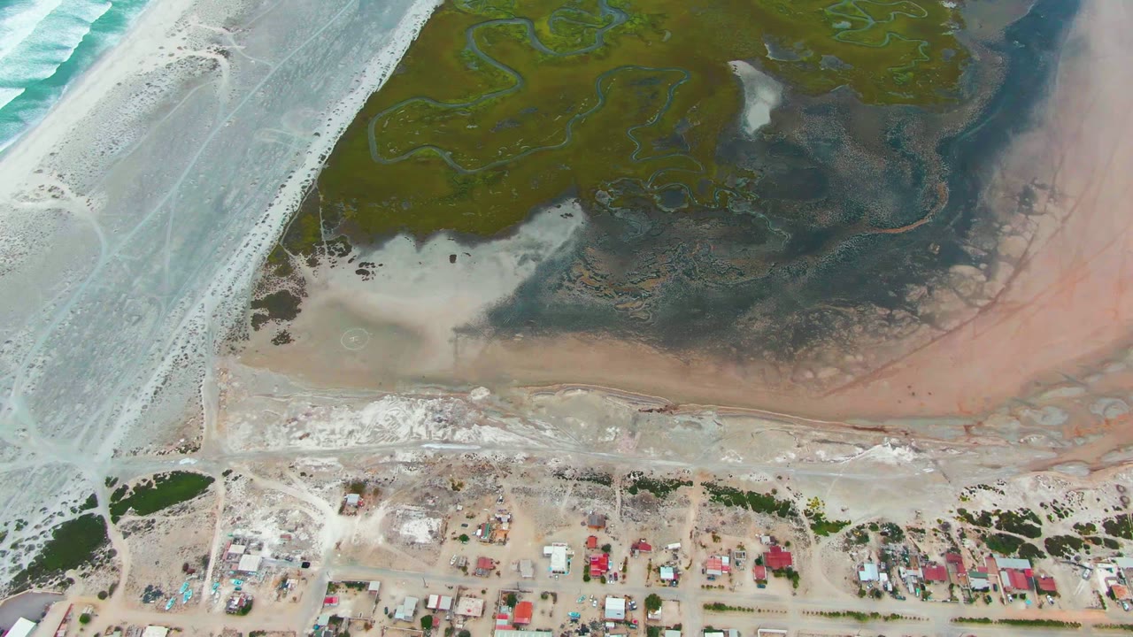 Overhead coastline with pale beach, green marsh, dark mudflat, and small settlement.
