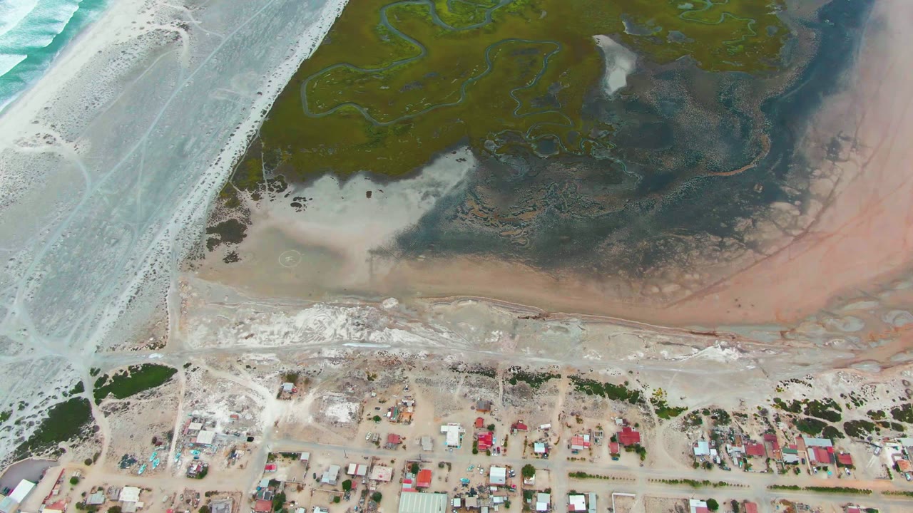 At 0:02, Punta Abreojos appears from above with turquoise-white surf on the left, green marsh and winding channels at top, pink-tan exposed flats along the shore, houses at bottom, and no visible people.