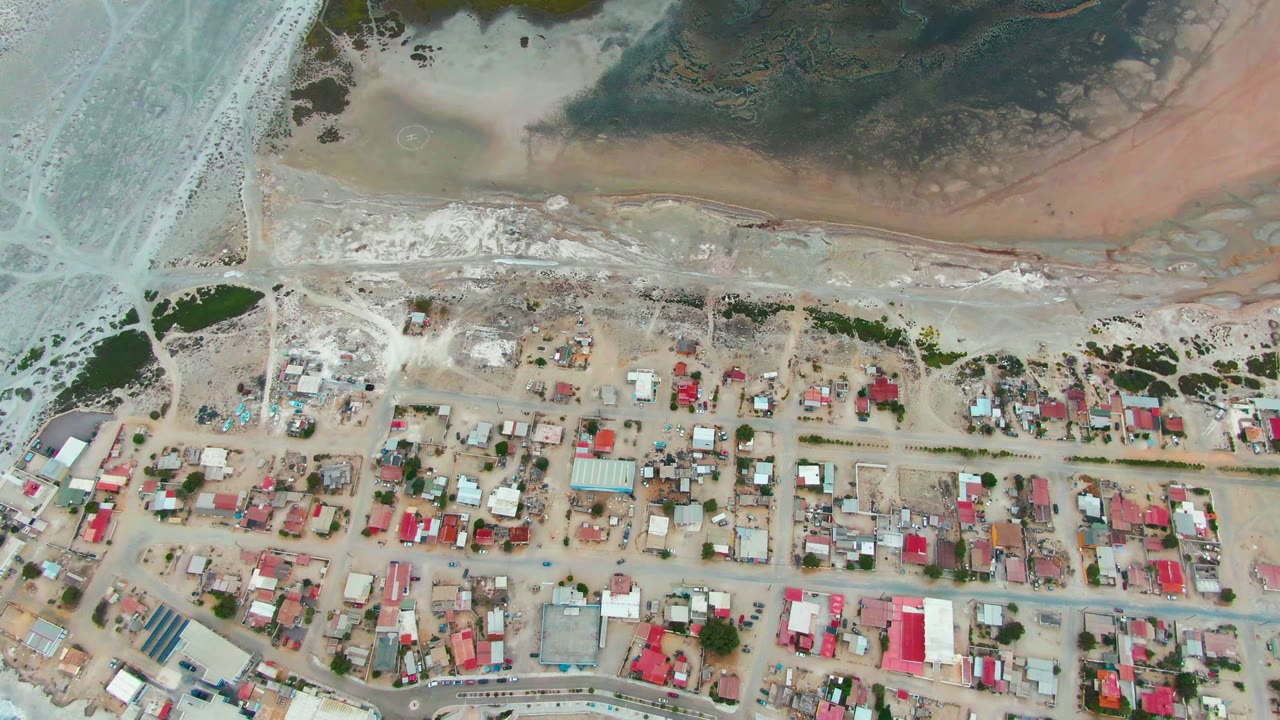 High aerial view of Punta Abreojos, Baja California Sur, with red-roofed houses, dirt streets, sandy shore, pale blue cove water, and no visible people.