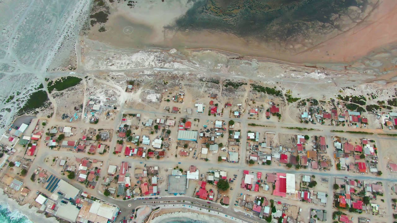 Top-down aerial of Punta Abreojos with red and pale roofs, sandy lots, a curving shoreline road, foamy surf, shallow cove water, and no visible people.