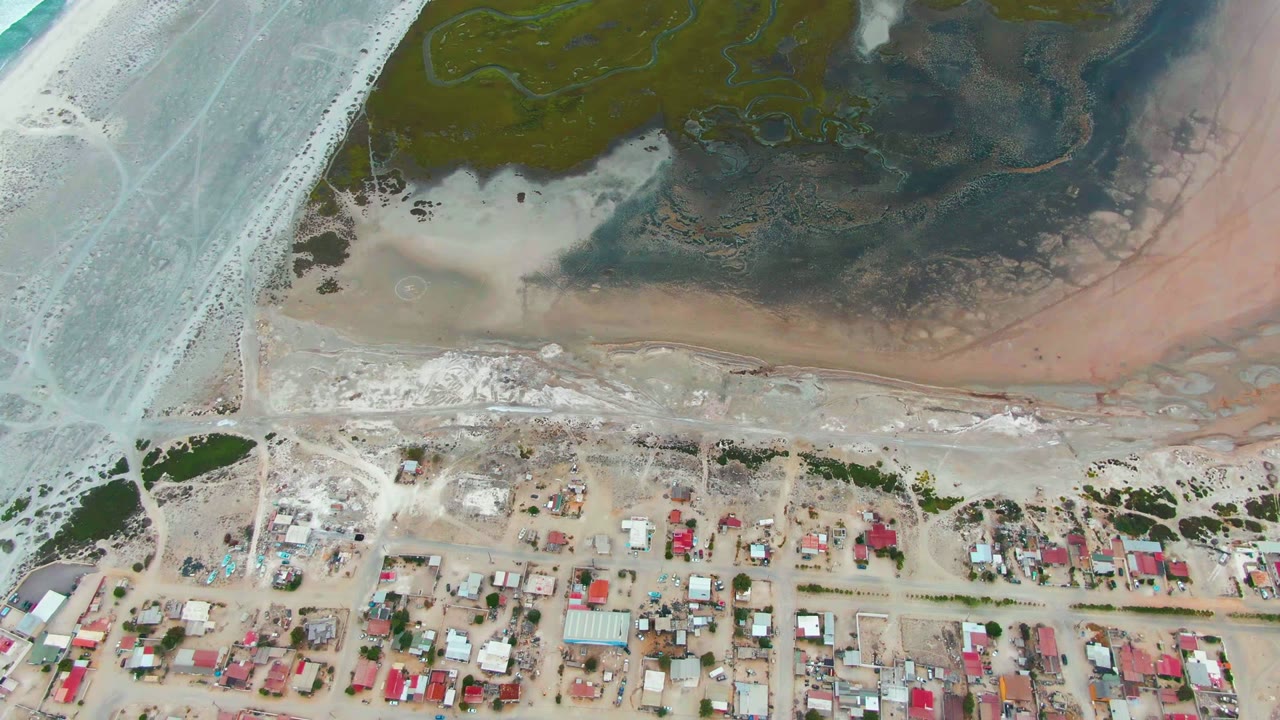 Top-down view of Punta Abreojos shoreline with pale surf at left, dark tidal flats above the beach, low homes below the road, and no visible people.