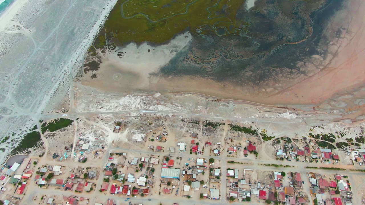 Aerial shoreline with wet sand, road, and clustered roofs.
