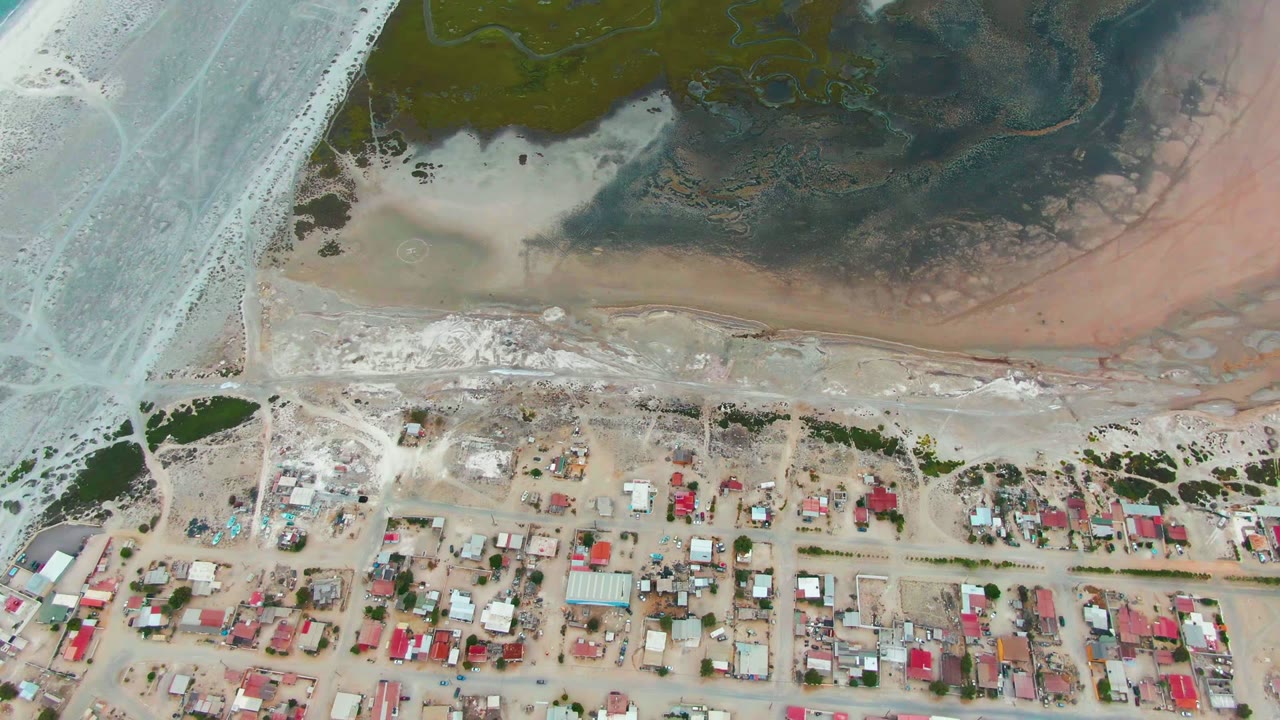 Aerial view over Punta Abreojos with pale blue water on the left, darker tidal shallows at top, sandy ground, residential blocks, and no visible people.