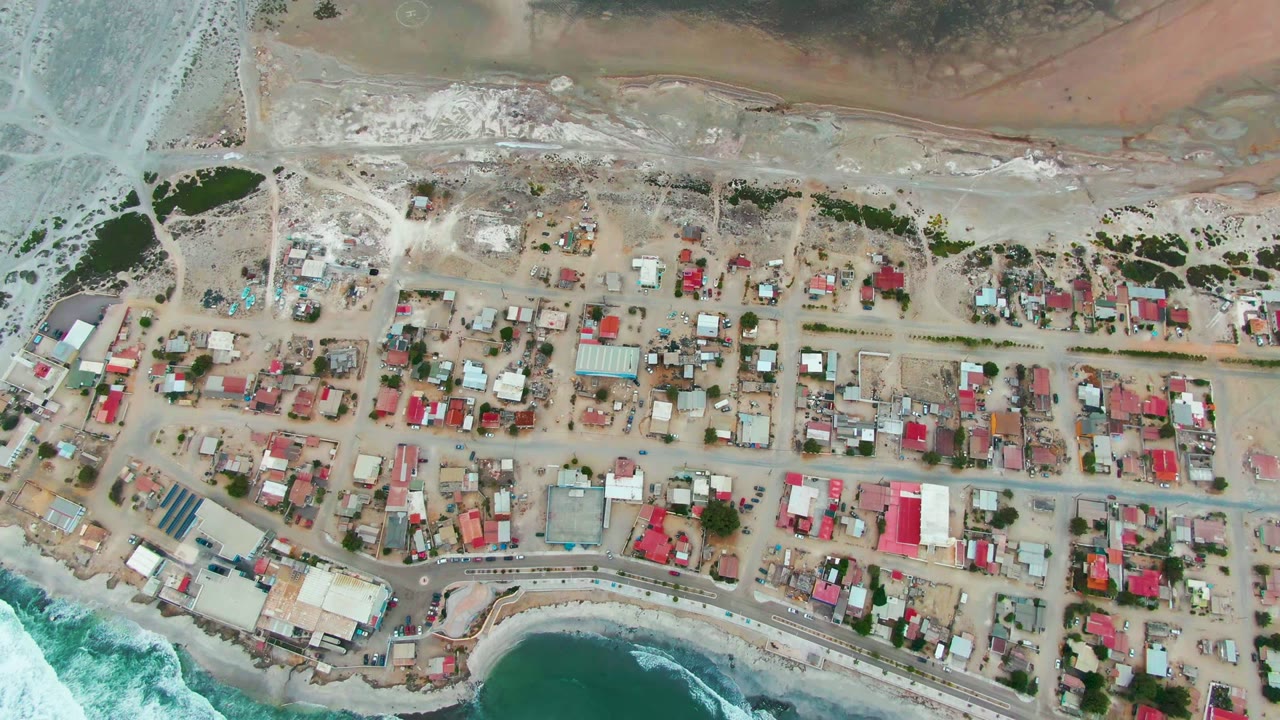 Top-down aerial frame of Punta Abreojos, Baja California Sur, Mexico, showing a small coastal village, gridded roads, pale sandy ground, surf at the lower edge, and no visible people.