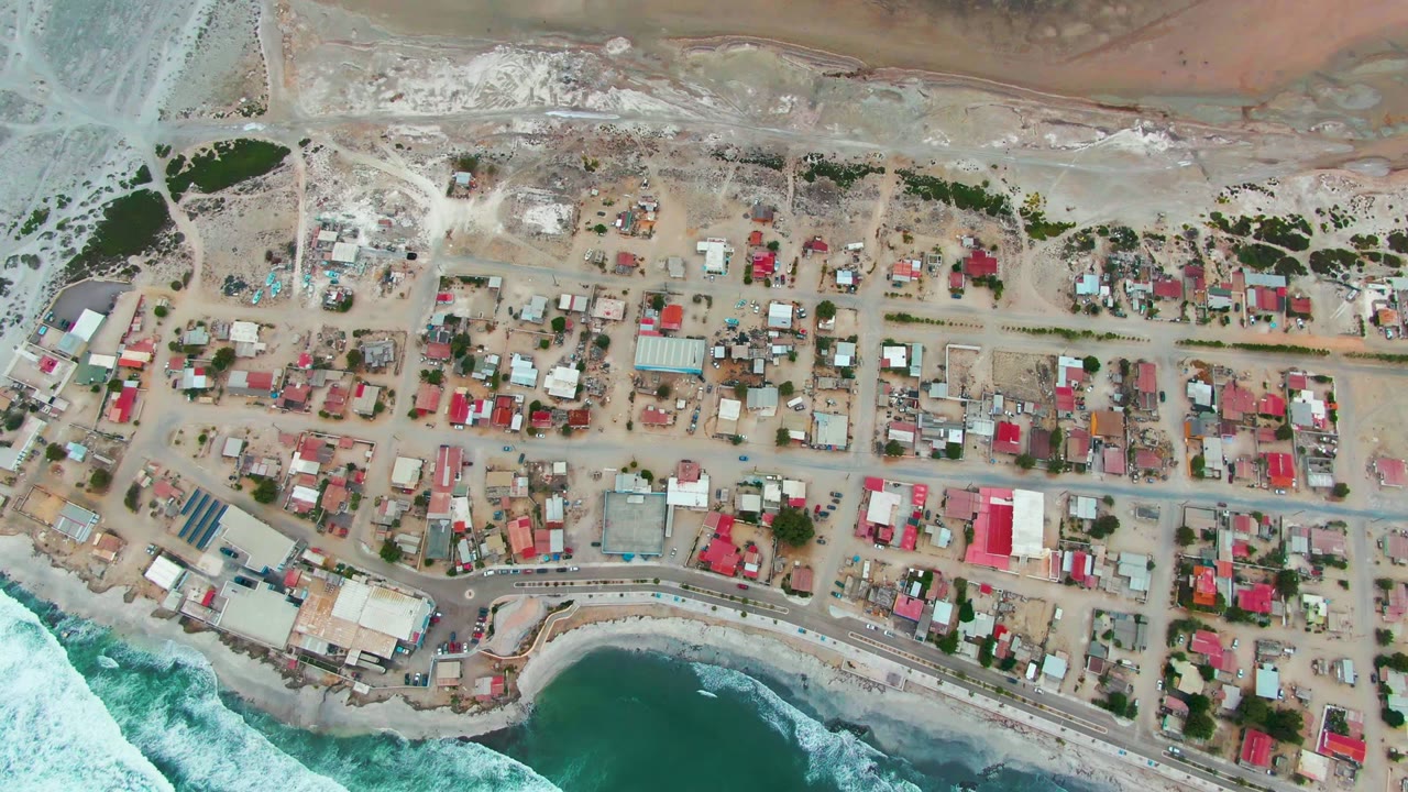 Overhead frame of the Punta Abreojos shoreline settlement, with dry pale flats above the houses, dense roofs and roads through the middle, ocean surf below, and no visible people.