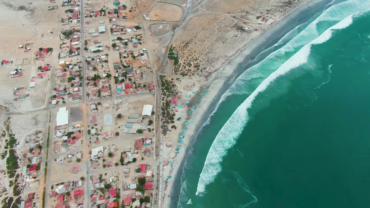 Overhead view of Punta Abreojos with town roads and small buildings on the left, sandy beach in the center, turquoise ocean on the right, and no visible people.
