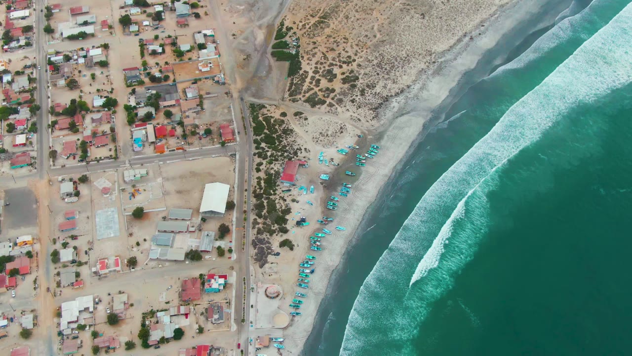 Overhead view of Punta Abreojos with tan residential blocks, a road, scrub dunes, blue beach umbrellas or boats, and turquoise waves breaking along shore, with no visible people.