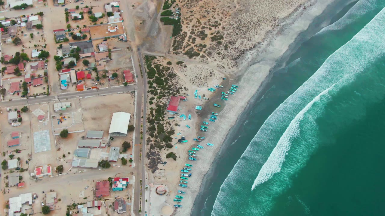 High drone view of Punta Abreojos with sandy lots and buildings on one side, beach scrub and a row of blue umbrellas or boats near shore, and an angled turquoise wave breaking offshore, with no visible people.