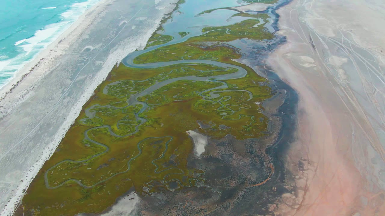 Overhead drone frame at Punta Abreojos, Baja California Sur showing turquoise surf on the left, pale beach, green delta channels, pink sediment flats, and no visible people.