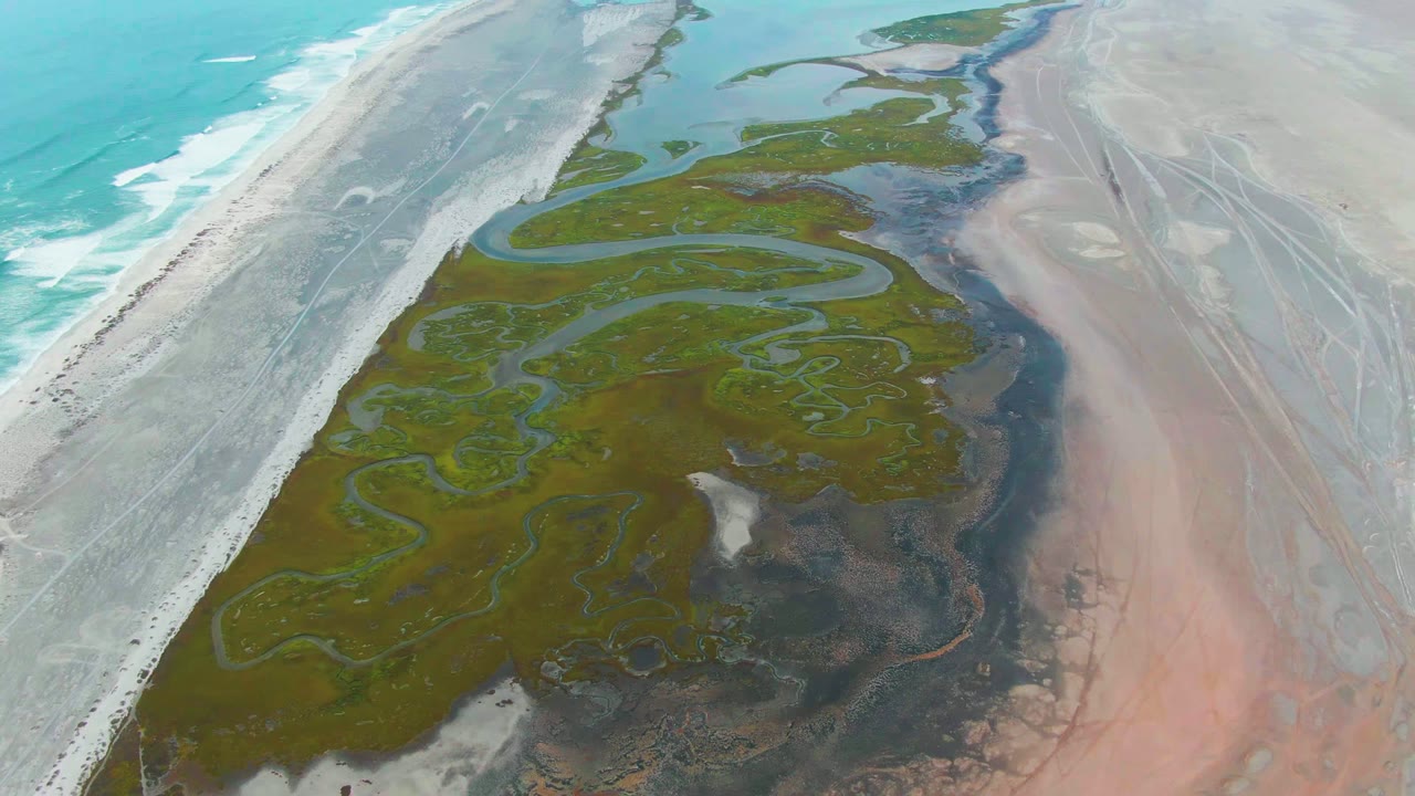 Aerial coastline with surf, beach, green delta, and shallow channels.