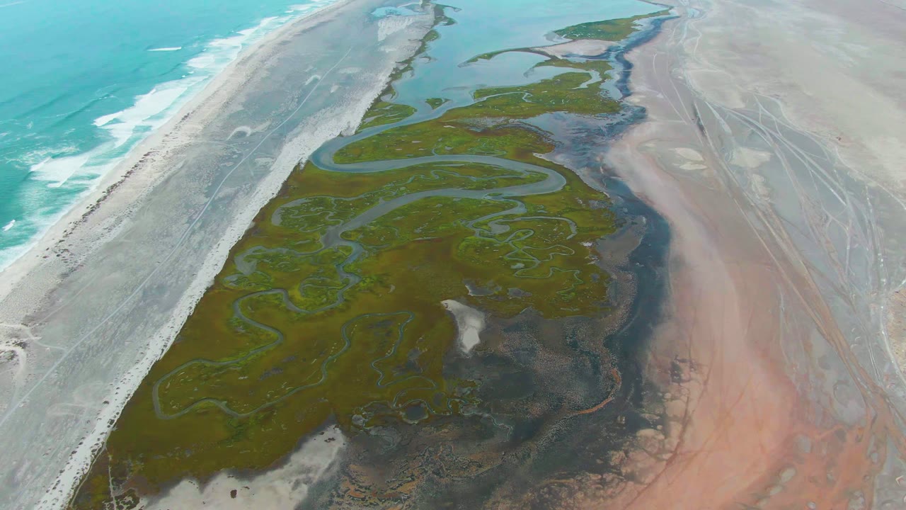 Mid-clip aerial frame in Punta Abreojos with ocean foam along the left edge, a broad green tidal delta in the center, pale sand flats to the right, and no visible people.