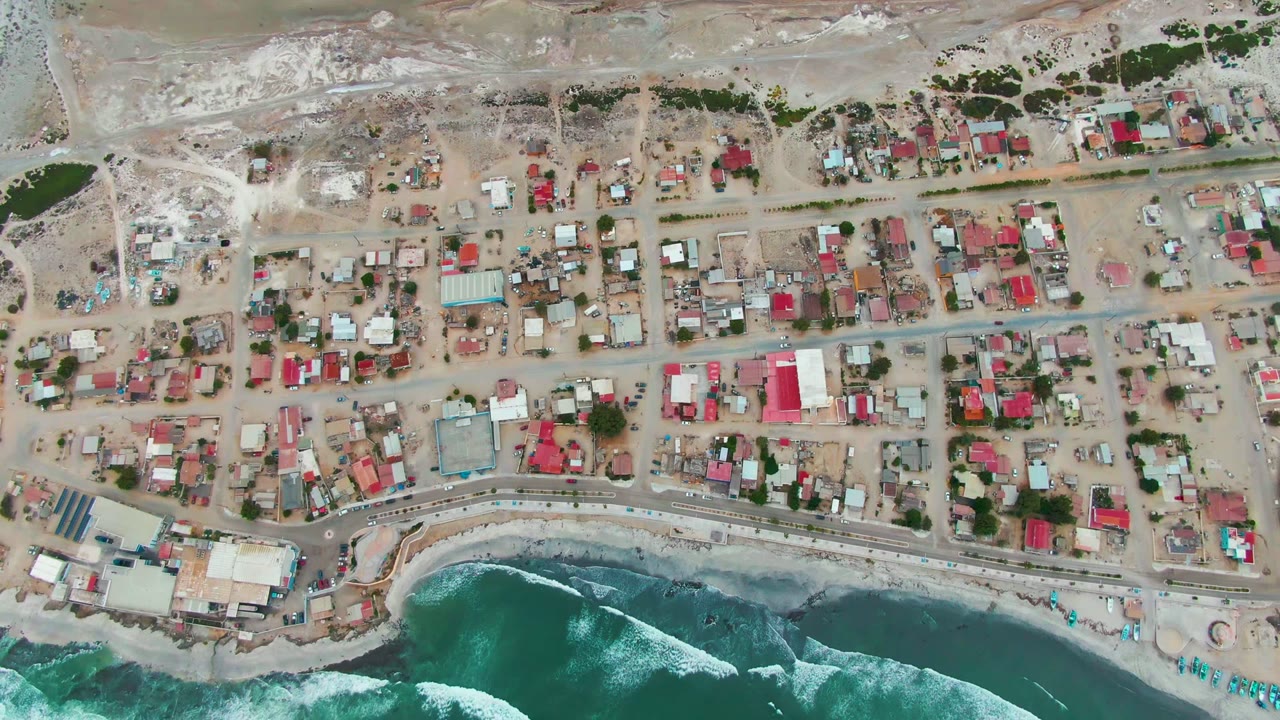Overhead view of Punta Abreojos with sandy streets, small houses, red roofs, a shore road, and blue-green waves breaking at the lower edge, with no visible people.