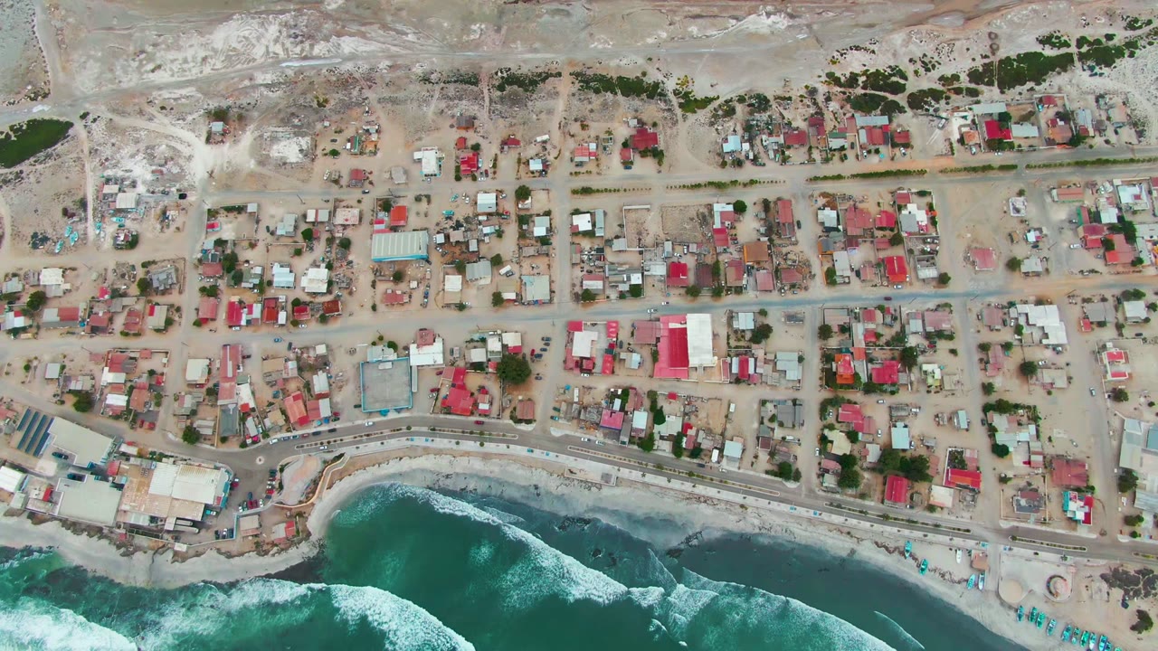 Top-down view of Punta Abreojos with rectangular houses and sandy roads above a narrow beach, while blue-green water and white surf fill the bottom of frame, with no visible people.