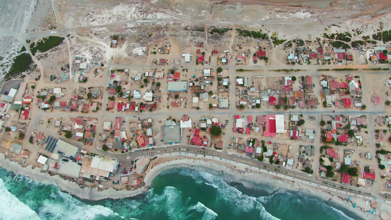 Overhead view of Punta Abreojos in Baja California Sur, with compact houses, sandy roads, dry ground inland, blue-green surf along the shore, and no visible people.