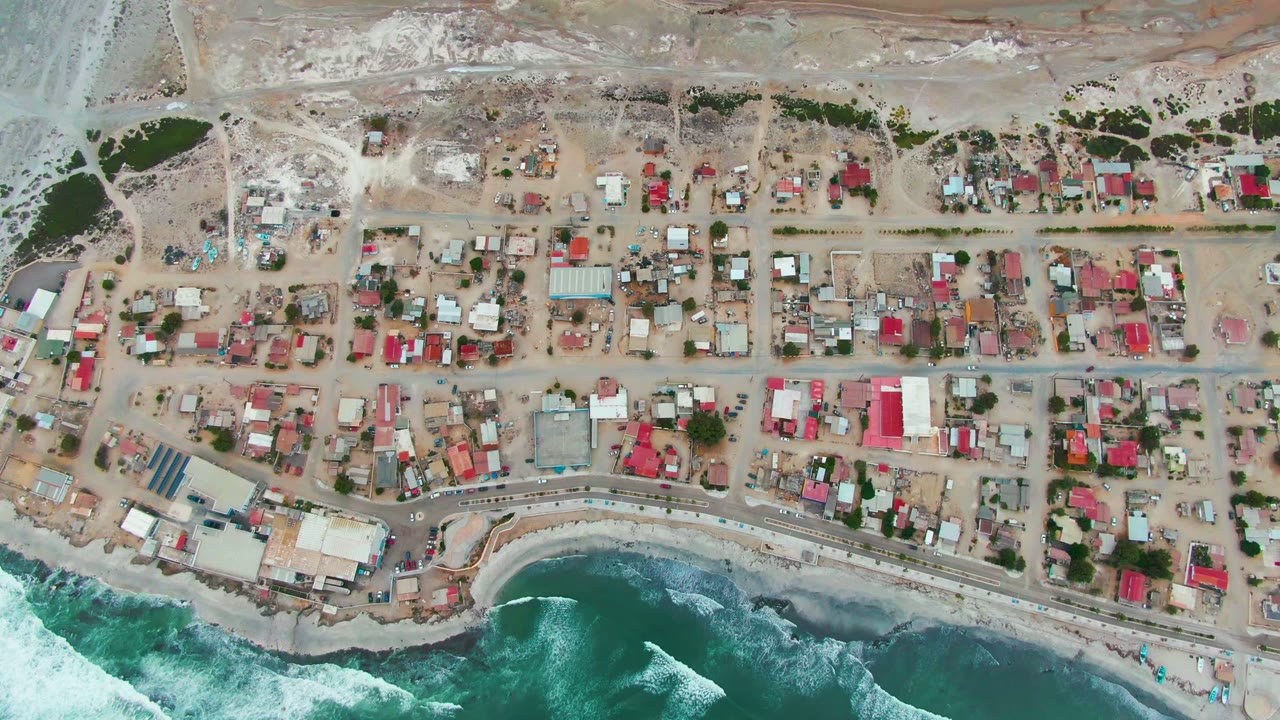 High overhead view of the Punta Abreojos shoreline, with rectangular town blocks, mixed roof colors, pale sand lots, waves curling below, and no visible people.