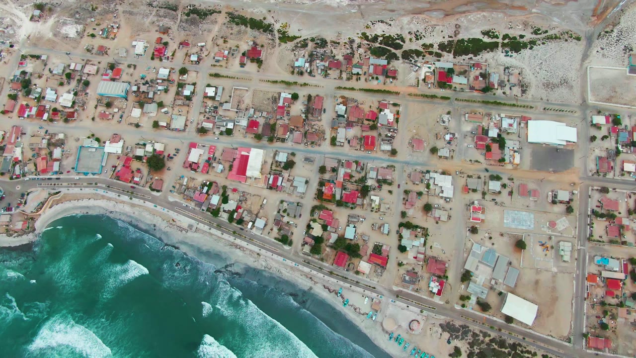 Overhead view of Punta Abreojos with turquoise surf at the bottom, a narrow sandy beach and shoreline road, and compact homes with red and white roofs inland.