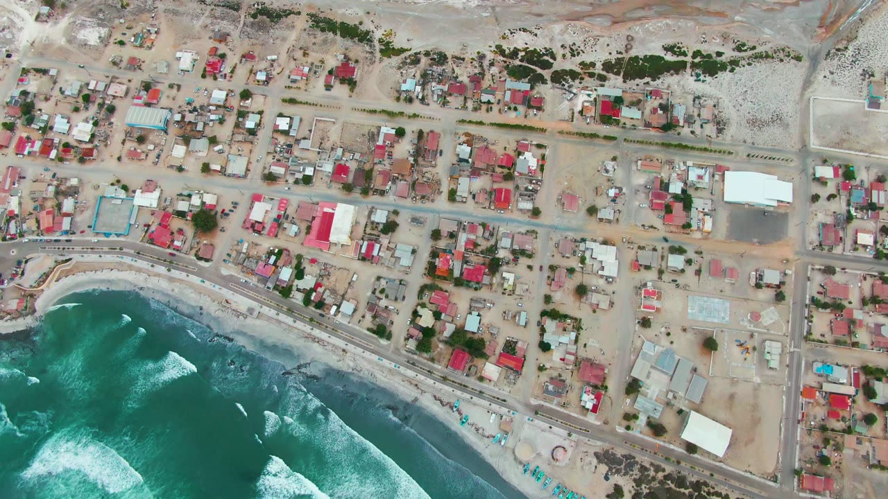 Aerial shoreline with waves, beach, road, and residential blocks.