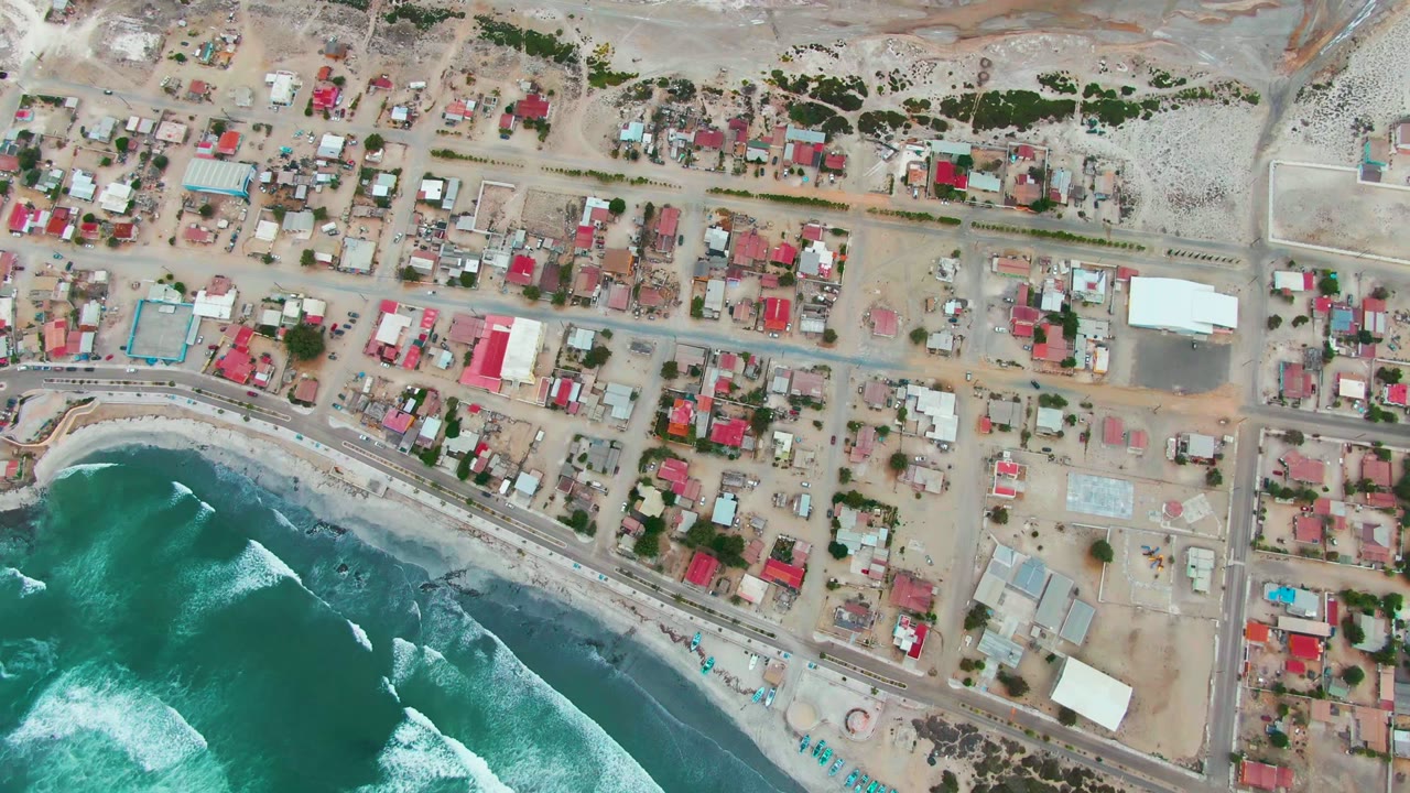 Top-down Punta Abreojos frame with a curved band of dark sand, turquoise water, white surf breaks, and rows of homes and dirt streets above the shore road.