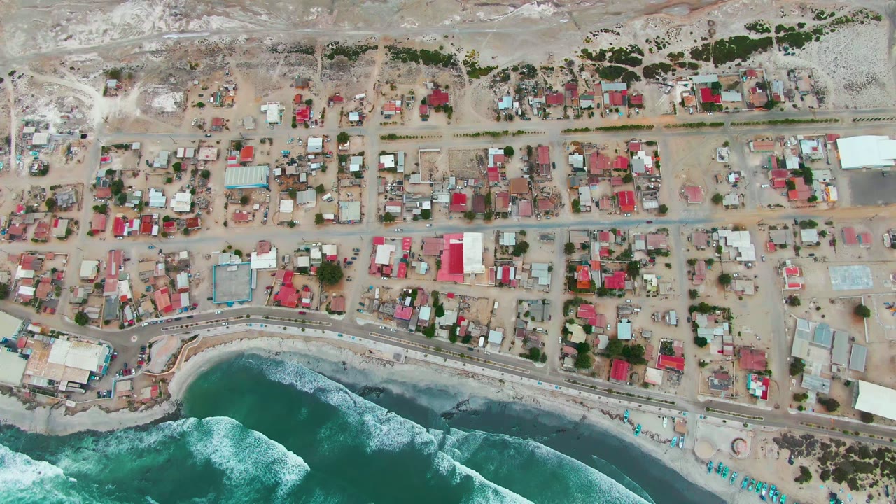 Overhead view of Punta Abreojos, Baja California Sur, with small homes, sandy streets, a shoreline road, turquoise surf, and no visible people.