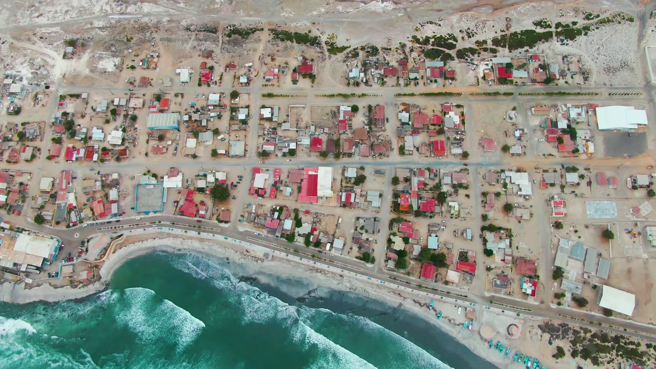 Top-down view of Punta Abreojos with compact residential blocks, a pale sandy shore, green-blue water, white breaking waves, and no visible people.