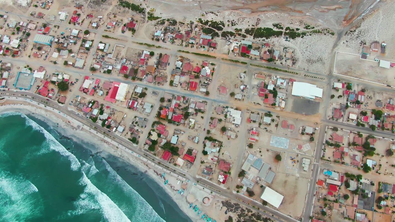 Top-down aerial view of Punta Abreojos with turquoise waves entering from the lower left, a sandy shore road, compact residential blocks, and no visible people.
