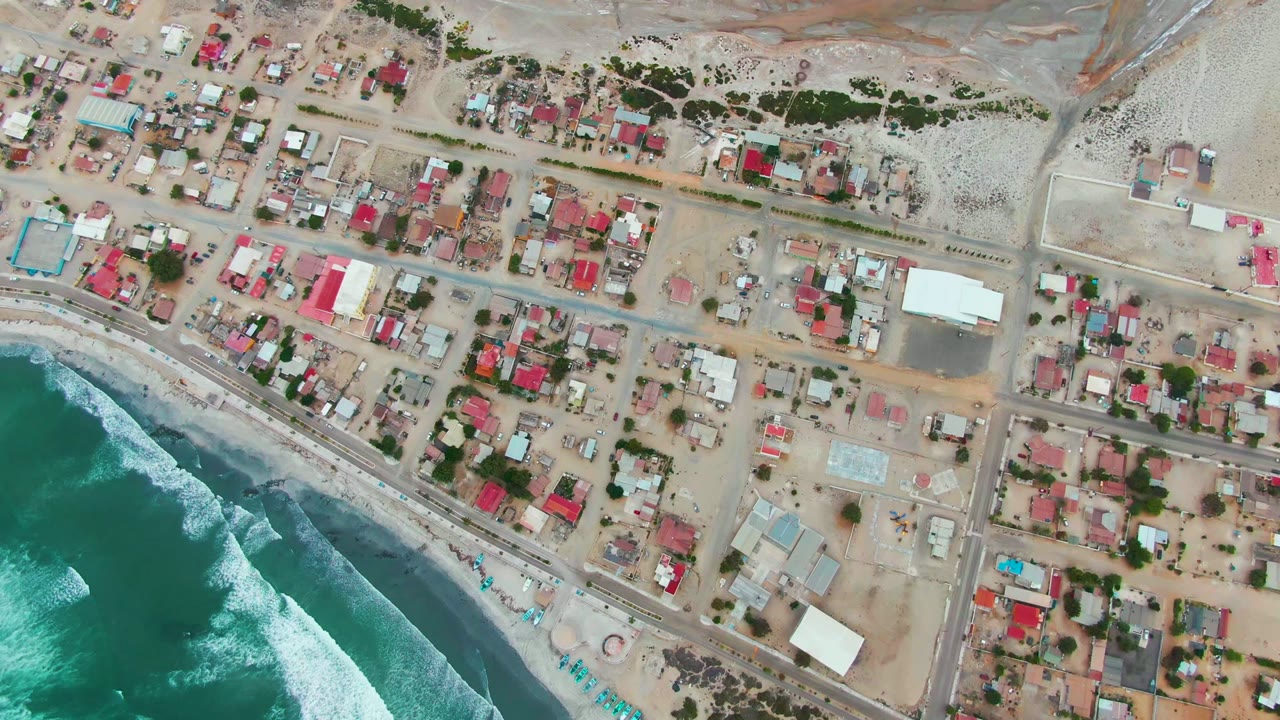 Mid-clip aerial frame over Punta Abreojos with foamy ocean water at the lower left, parallel shore structures, grid streets, scattered rooftops, and no visible people.