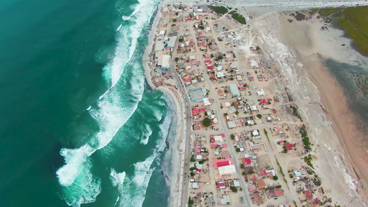 Aerial frame over Punta Abreojos, Baja California Sur, with turquoise ocean on the left, white surf near shore, small buildings inland, and no visible people.