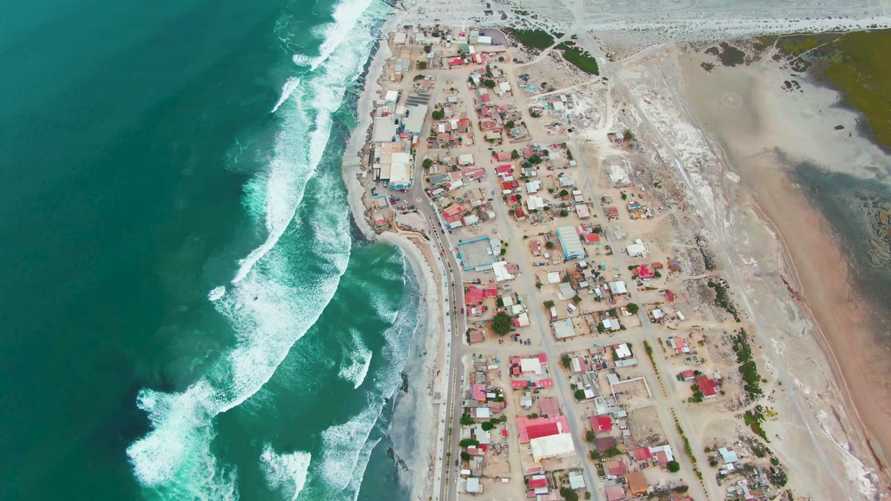 Aerial frame of Punta Abreojos showing foamy breakers on the left, a dense cluster of small roofs near the center, open sand to the right, and no visible people.
