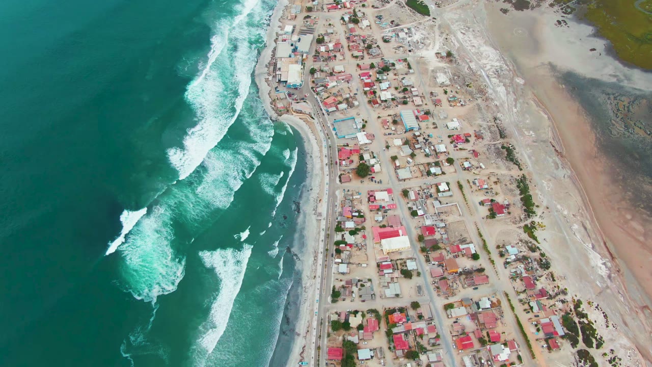 High aerial frame over Punta Abreojos shows turquoise ocean on the left, white waves approaching the sandy beach, shoreline roads, low buildings, and no visible people.