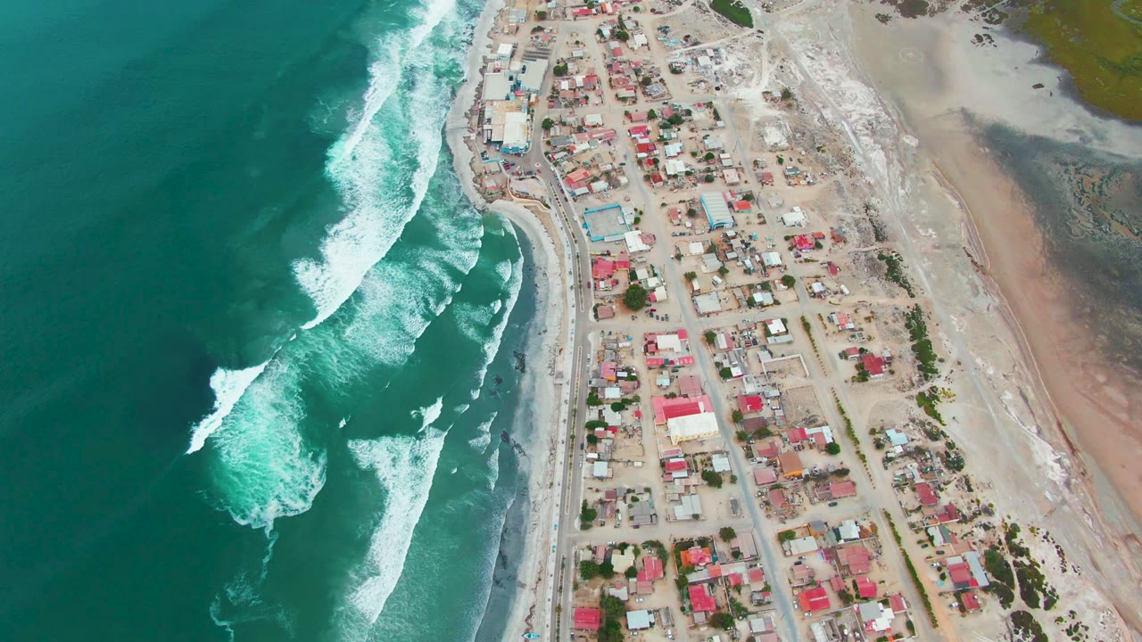 Breaking ocean waves run parallel to a sandy beach and village roads.