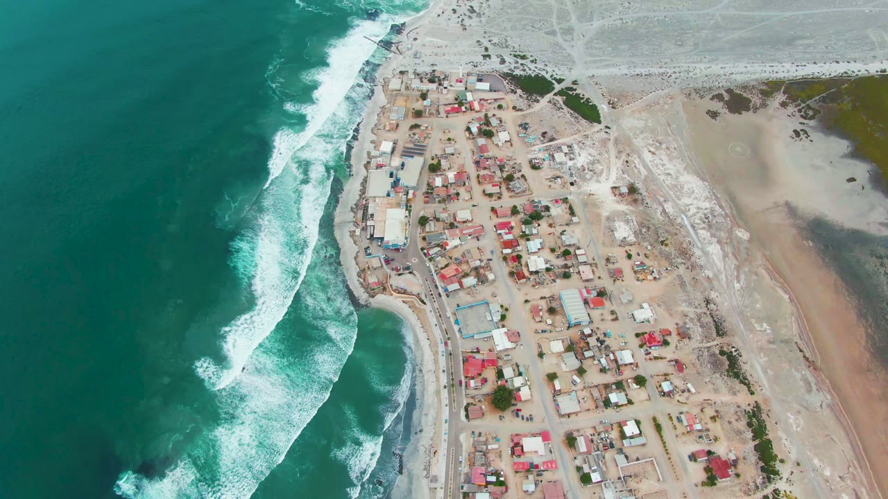 Opening aerial frame over Punta Abreojos, Baja California Sur, with turquoise ocean and white surf on the left, clustered buildings on sandy ground, pale terrain to the right, and no visible people.