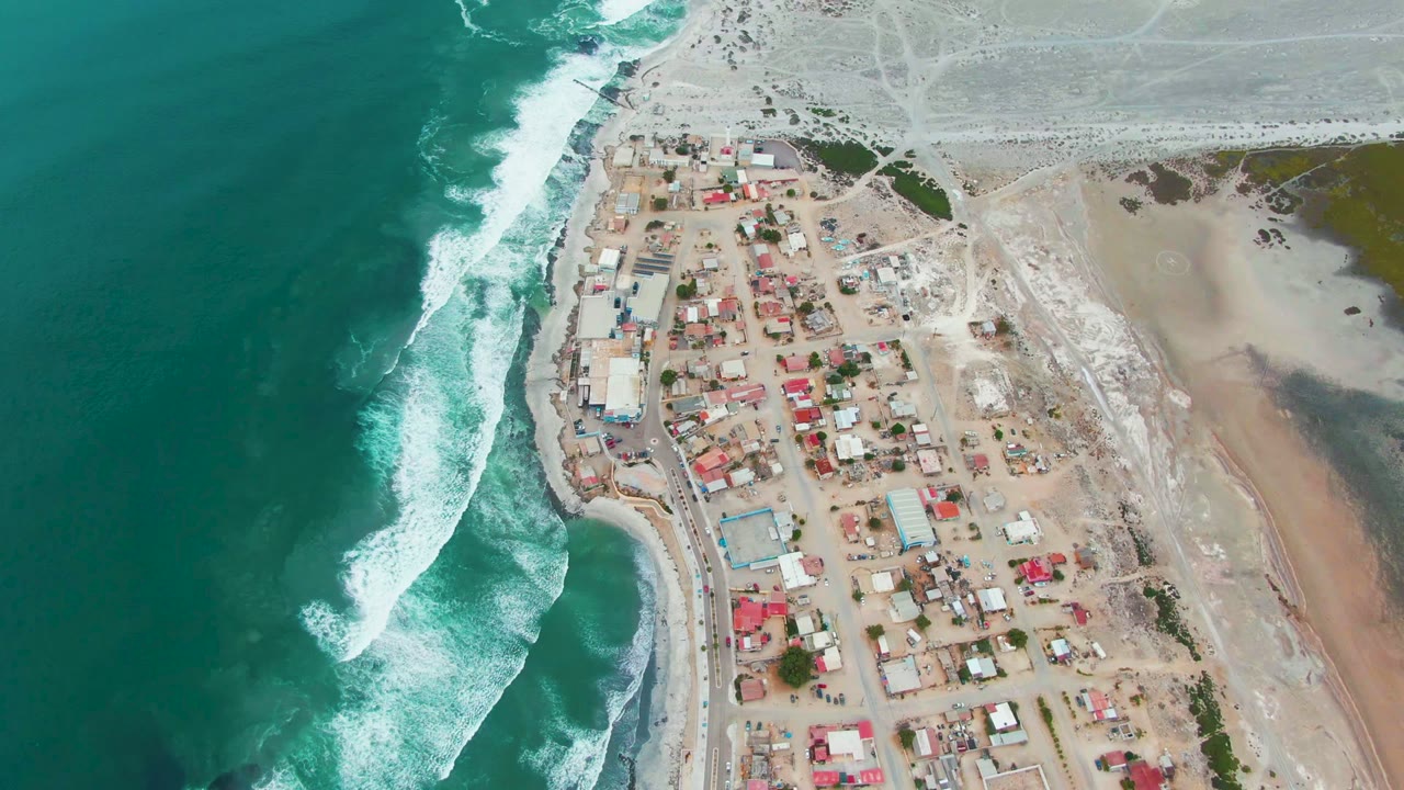Drone view of white surf rolling beside red-roofed buildings on sand.