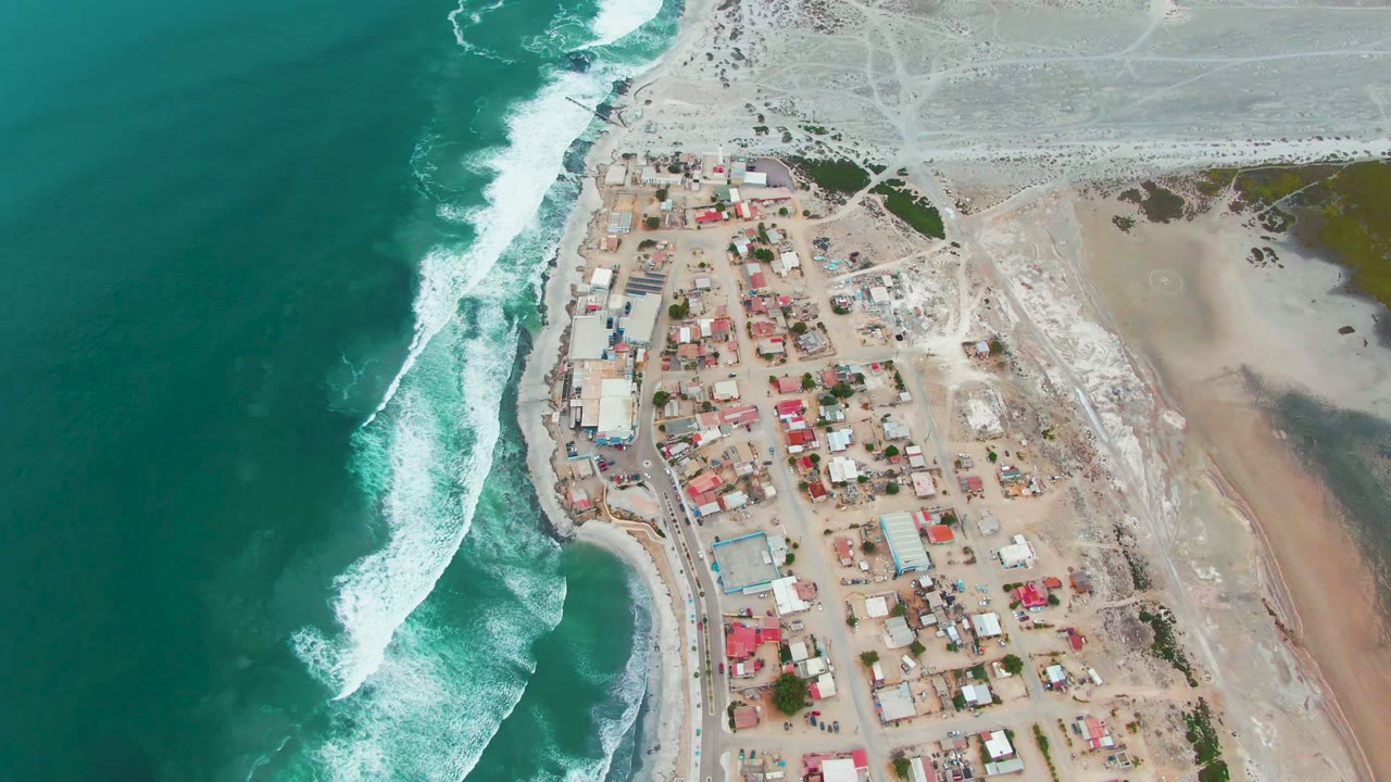 Drone frame over Punta Abreojos with foamy waves advancing along the left edge, a compact block of low buildings near the shore, open pale terrain inland, and no visible people.
