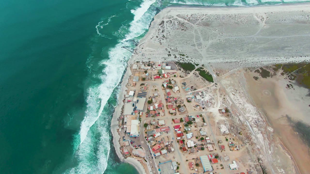 Aerial frame over Punta Abreojos, Baja California Sur, with turquoise ocean on the left, white surf at the shore, clustered small buildings below, and no visible people.