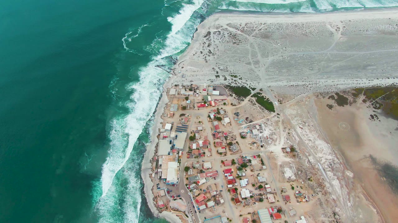 High overhead coastal settlement beside breaking waves and pale sandy flats.