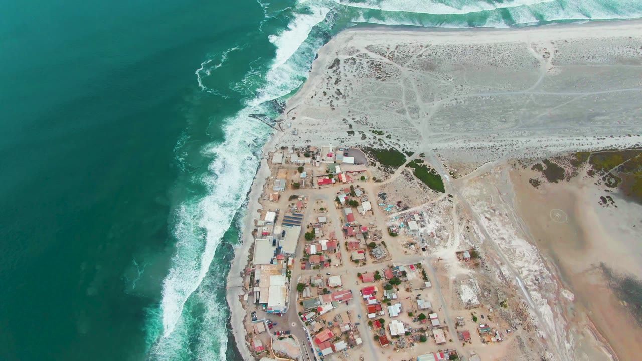 Aerial view in Punta Abreojos, Baja California Sur, showing foamy waves along the left shoreline, small buildings near the lower center, pale sand above, and no visible people.