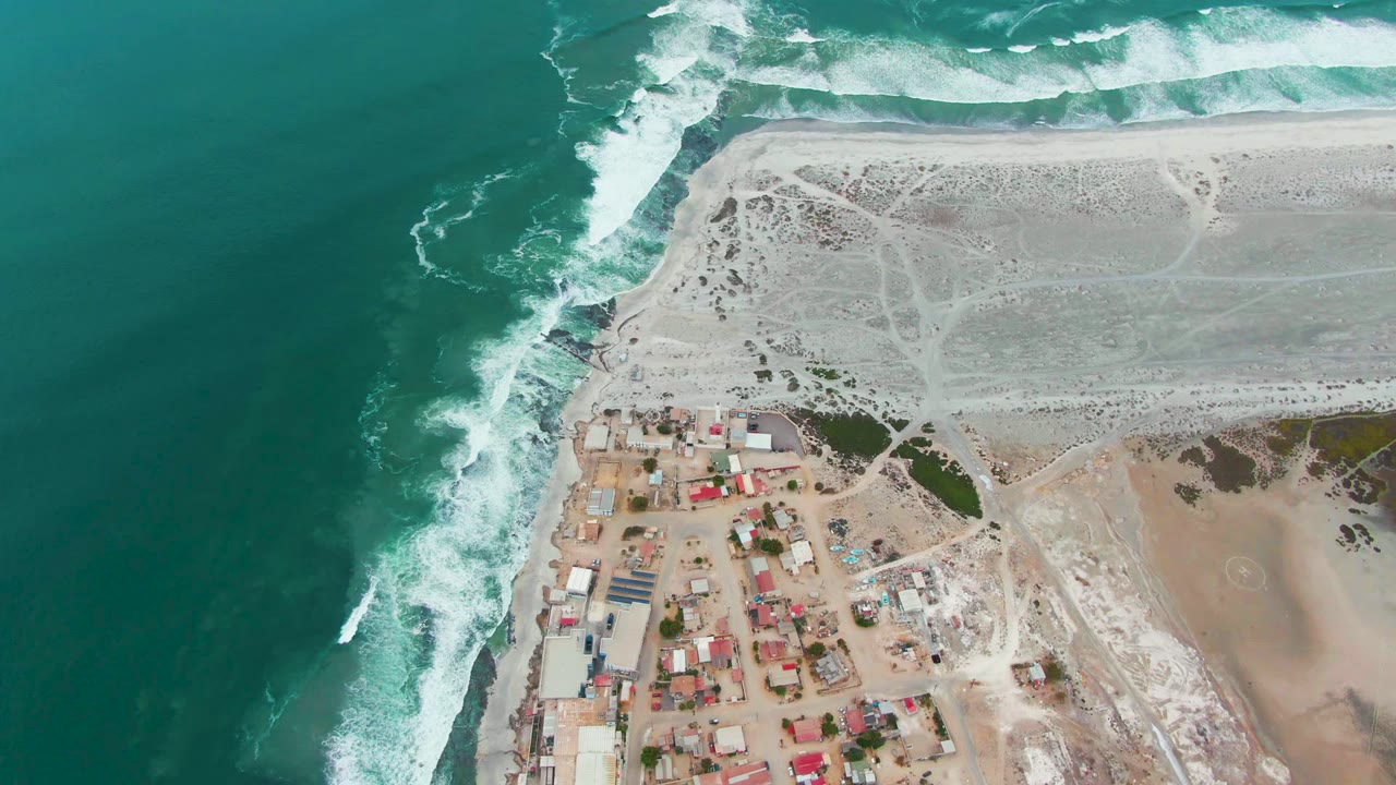 Opening aerial frame over Punta Abreojos, Baja California Sur, with turquoise water left, white waves breaking along the shore, clustered buildings below, and no visible people.