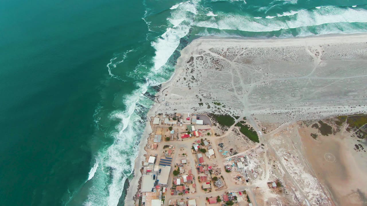 Overhead coastline with surf wrapping around a rocky point and buildings near the beach.