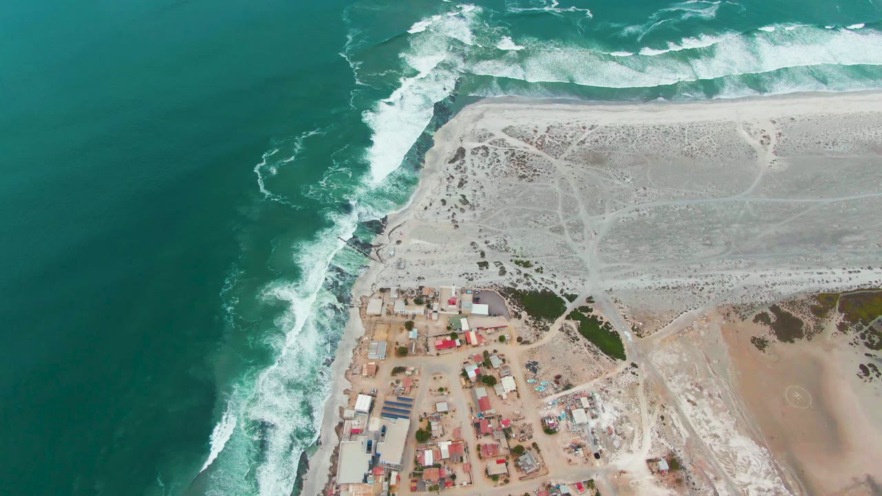 The frame holds an overhead composition of Punta Abreojos with dense white surf at the top shore, deep turquoise water on the left, scattered buildings below, and no visible people.