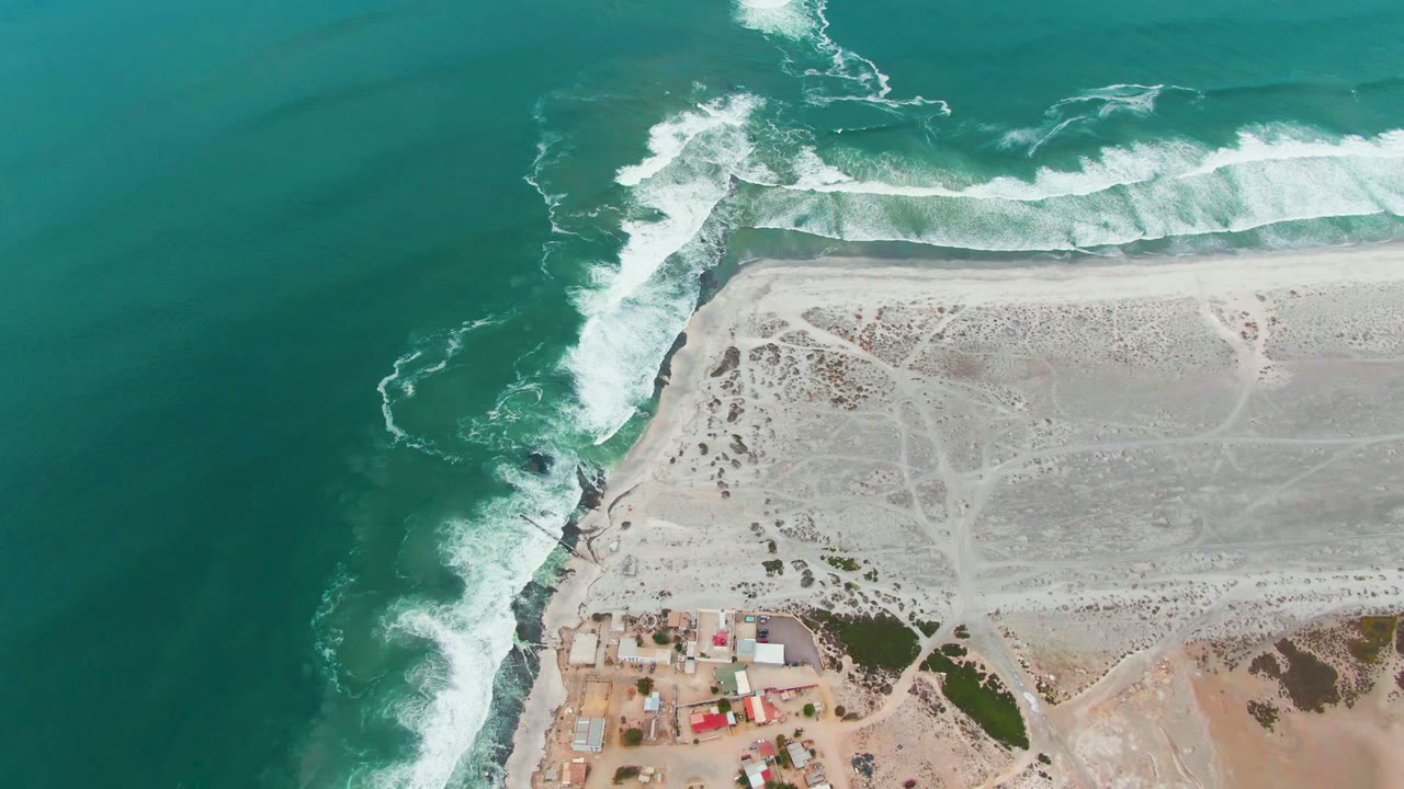 Opening overhead frame in Punta Abreojos, Baja California Sur showing turquoise water, white surf wrapping a rocky point, pale sand, small buildings at the lower edge, and no visible people.
