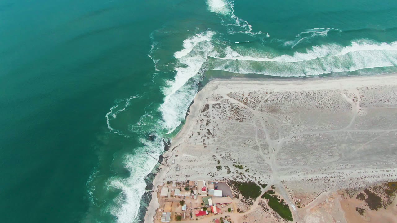 Overhead coastline with whitecaps rolling into a rocky point beside sandy land.