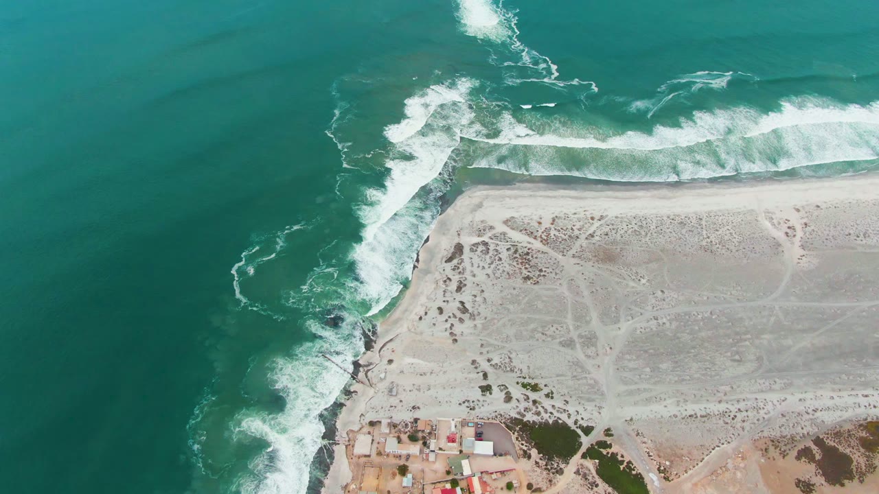 At 0:02, Punta Abreojos coastline appears from above with a wide turquoise sea field, foamy waves along the top beach, rocks at the bend, inland sand tracks, small buildings, and no visible people.