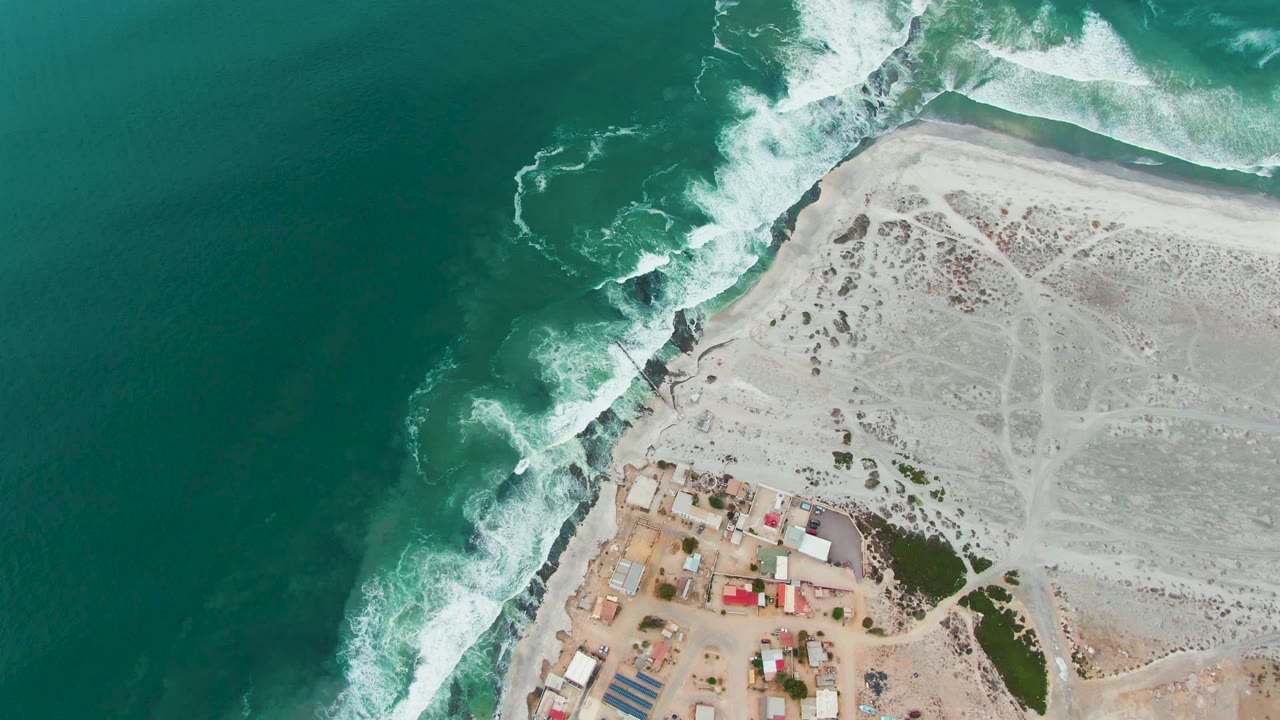 Drone view of waves curling onto a rocky shore beside sand and a small coastal settlement.