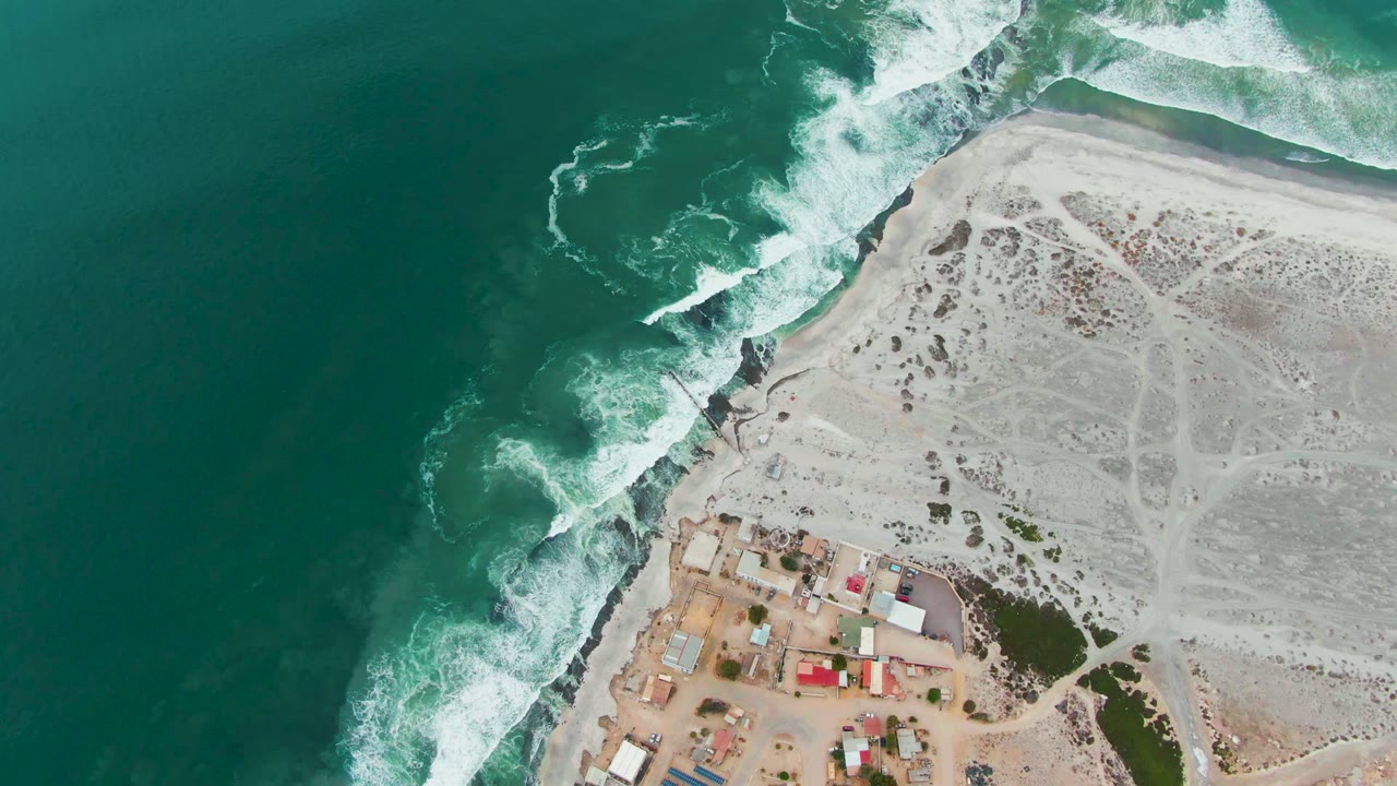At 0:02, a broader patch of green ocean fills the left side while white breakers trace the Punta Abreojos coast, with sandy ground and small structures on the lower right and no visible people.