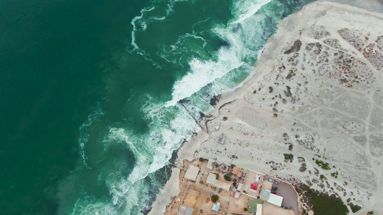 Overhead view of Punta Abreojos surf breaking into white foam beside pale sand, dark shoreline rocks, and low coastal buildings near the bottom edge, with no visible people.