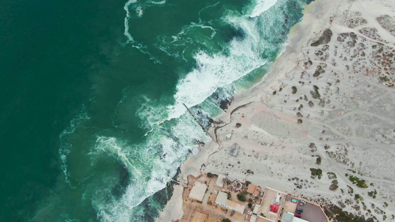 Overhead Punta Abreojos coastline with foamy surf curling around dark rocks, open sand marked by sparse scrub, and small buildings partly visible at the bottom, with no visible people.