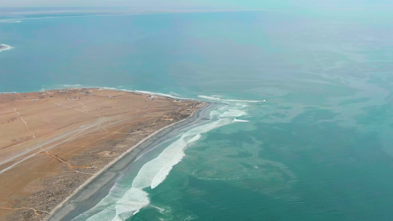 High aerial view of Punta Abreojos coastline with tan land at left, blue-green ocean at right, white waves along a dark beach, and no visible people.