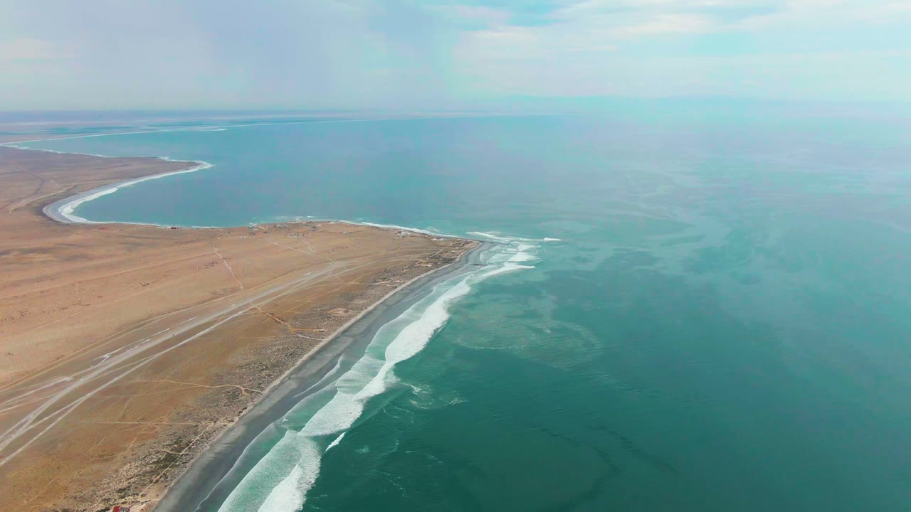 Opening aerial view of Punta Abreojos, Baja California Sur, with tan desert land, a shoreline road, white surf bands, blue ocean, scattered clouds, and no visible people.