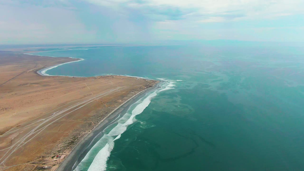 Punta Abreojos coastline seen from above, with a curved bay in the distance, tan desert terrain, a narrow road, white wave lines, turquoise ocean, and no visible people.