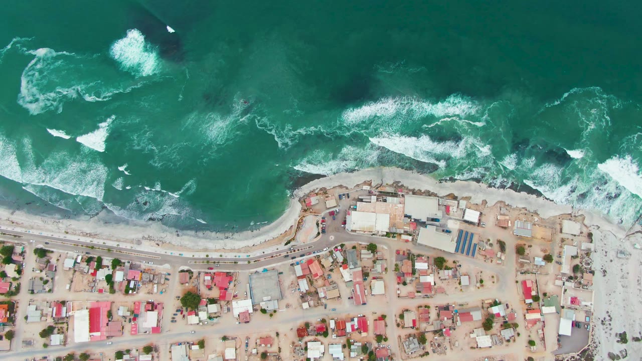 Top-down aerial view of Punta Abreojos, Baja California Sur, with green ocean surf above a sandy beach and compact village buildings below, no visible people.